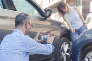Driver photographing vehicle damage at the scene of a crash while another person calls for help